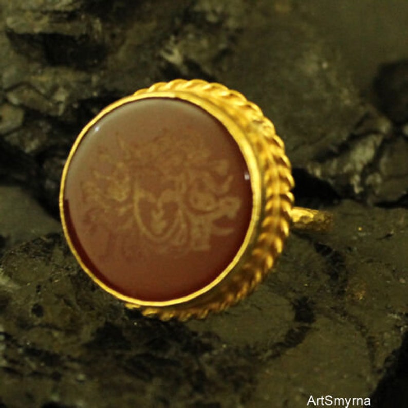 May include: A gold-colored ring with a round, reddish-brown stone. The stone has a detailed, light-colored design etched into its surface. The ring's band has a textured, braided design around the stone.