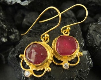 May include: Gold earrings with a red gemstone and a small clear stone accent. The earrings have a delicate, ornate design.