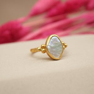 May include: A gold ring with a white pearl-like stone set in a textured gold setting. The ring is accented with small gold beads.