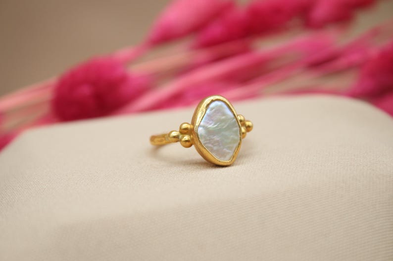May include: A gold ring with a white pearl-like stone set in a textured gold setting. The ring is accented with small gold beads.