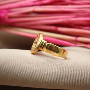 Op de afbeelding: Een gouden ring met een getextureerd ontwerp. De ring ligt op een beige stoffen achtergrond.