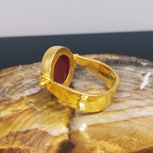 Peut inclure: Une bague en or avec une pierre pr&eacute;cieuse rouge sertie dans une b&eacute;li&egrave;re ronde. La bague a une finition martel&eacute;e et un design pivotant.