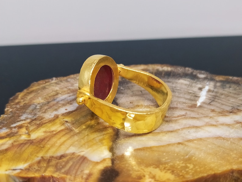 Peut inclure: Une bague en or avec une pierre pr&eacute;cieuse rouge sertie dans une b&eacute;li&egrave;re ronde. La bague a une finition martel&eacute;e et un design pivotant.