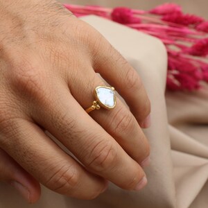 May include: A gold ring with a white, irregular shaped gemstone. The ring is on a finger.