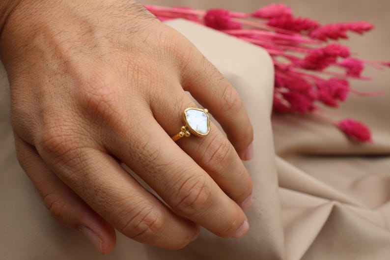 May include: A gold ring with a white, irregular shaped gemstone. The ring is on a finger.