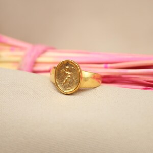 May include: A gold ring with an oval-shaped design featuring a raised image of a person. The ring is on a light pink and beige background.