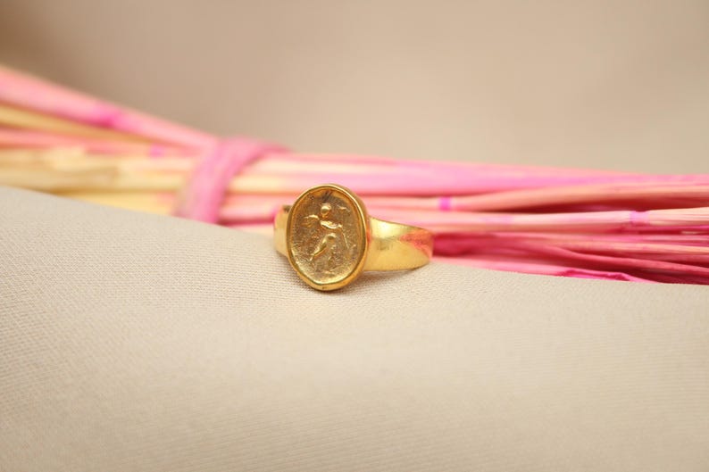 May include: A gold ring with an oval-shaped design featuring a raised image of a person. The ring is on a light pink and beige background.