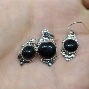 K&ouml;nnte beinhalten: Ein Set aus silbernen Ohrringen und einem Anh&auml;nger mit schwarzen Edelsteinen. Die Ohrringe und der Anh&auml;nger haben ein detailliertes Design mit einer silbernen Filigranarbeit.