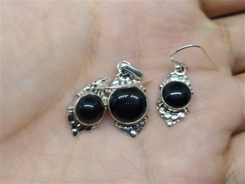K&ouml;nnte beinhalten: Ein Set aus silbernen Ohrringen und einem Anh&auml;nger mit schwarzen Edelsteinen. Die Ohrringe und der Anh&auml;nger haben ein detailliertes Design mit einer silbernen Filigranarbeit.