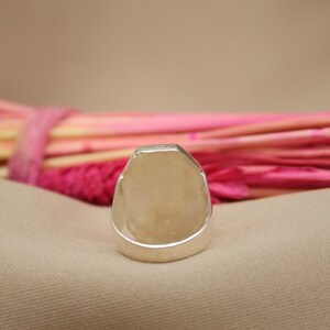 May include: A silver ring with a rectangular face. The ring is set against a neutral background with pink and yellow decorative elements. The ring's design is simple and modern, suitable for everyday wear.