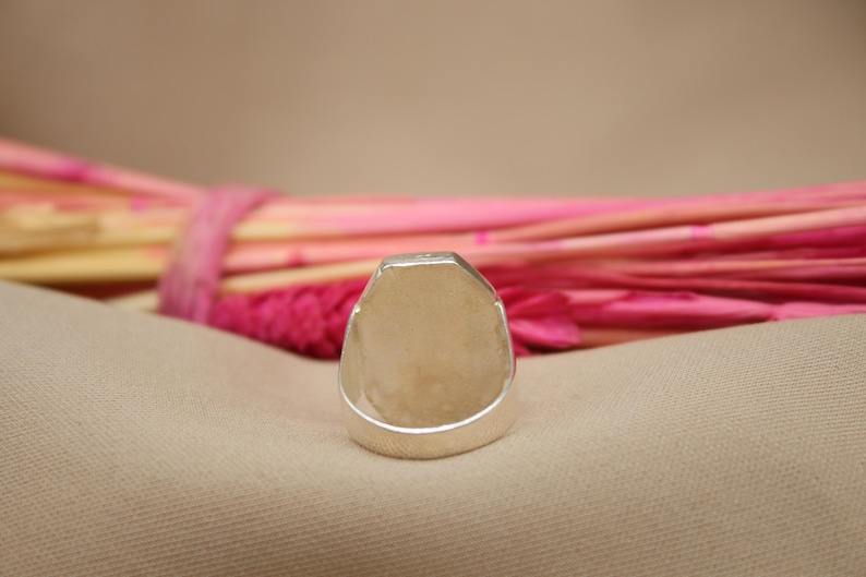 May include: A silver ring with a rectangular face. The ring is set against a neutral background with pink and yellow decorative elements. The ring's design is simple and modern, suitable for everyday wear.