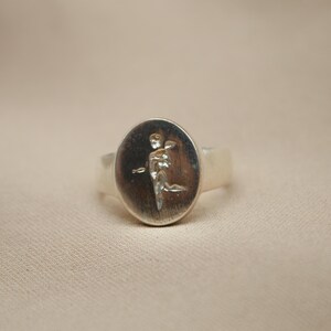 以下が含まれることがあります： 羽根のある人物のレリーフデザインが施された、楕円形のセッティングのシルバーリング。