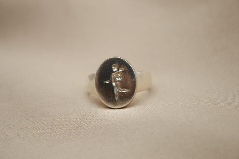 以下が含まれることがあります： 羽根のある人物のレリーフデザインが施された、楕円形のセッティングのシルバーリング。