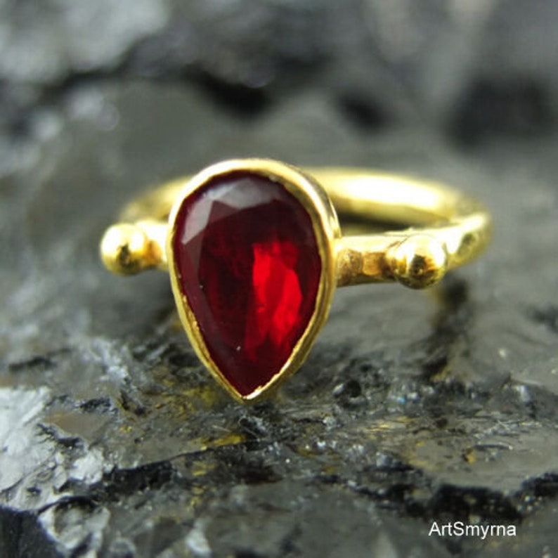 May include: A gold ring with a teardrop-shaped red gemstone. The ring has two small gold balls on either side of the gemstone.