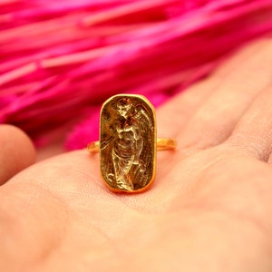 May include: A gold ring with a detailed design of a winged figure. The ring is on a hand against a pink background.