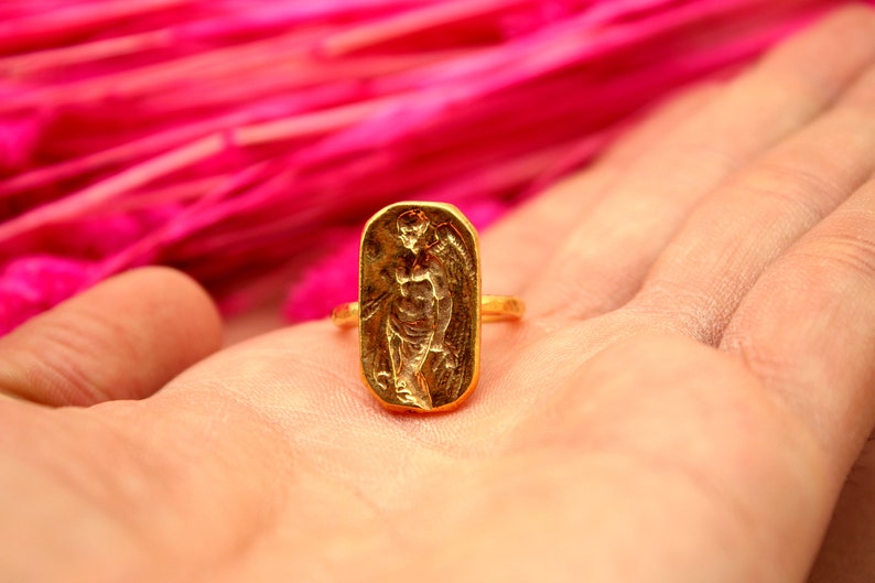 May include: A gold ring with a detailed design of a winged figure. The ring is on a hand against a pink background.