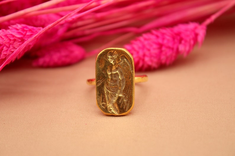 May include: A gold ring with a rectangular design featuring an embossed image of an angel with wings.