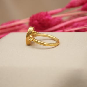 May include: A gold ring with a textured band and a round, faceted gemstone. The ring is set on a white surface.