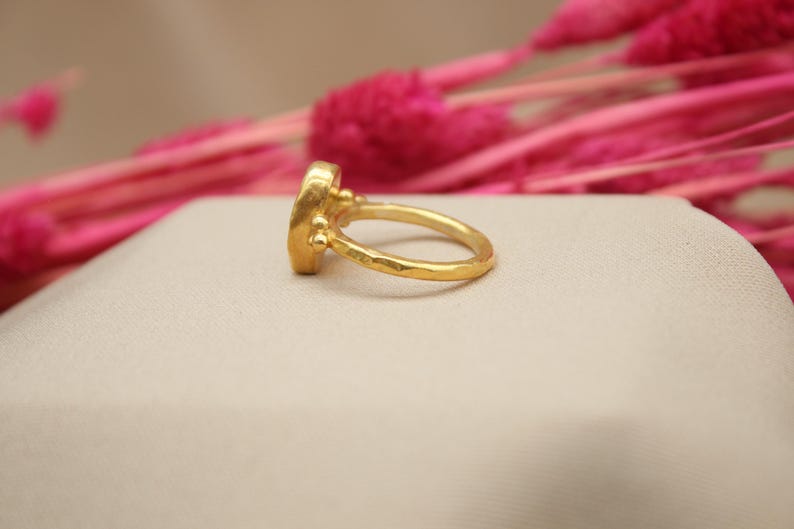 May include: A gold ring with a textured band and a round, faceted gemstone. The ring is set on a white surface.