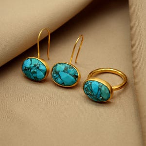 May include: A set of gold earrings and a ring with turquoise stones. The earrings are oval shaped and dangle from a gold hook. The ring is a simple band with a turquoise stone set in the center.