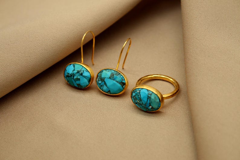 May include: A set of gold earrings and a ring with turquoise stones. The earrings are oval shaped and dangle from a gold hook. The ring is a simple band with a turquoise stone set in the center.