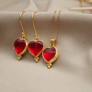 May include: A set of gold heart-shaped earrings and a necklace with red glass stones. The jewellery is on a beige fabric background.