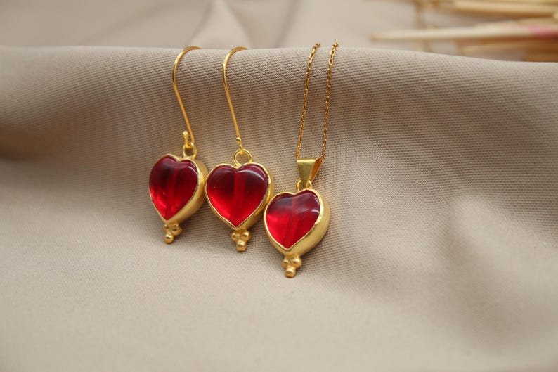 May include: A set of gold heart-shaped earrings and a necklace with red glass stones. The jewellery is on a beige fabric background.