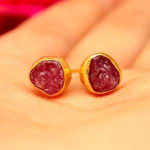 May include: A pair of stud earrings featuring raw, deep pinkish-red stones set in gold-toned metal bezels. The stones have an irregular, textured surface and are held in place by smooth, rounded gold-colored frames with visible earring posts.