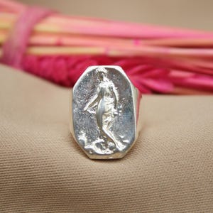 May include: A silver ring with a rectangular face featuring a sculpted figure. The ring is set against a neutral background with a blurred arrangement of pink and yellow stems in the background.