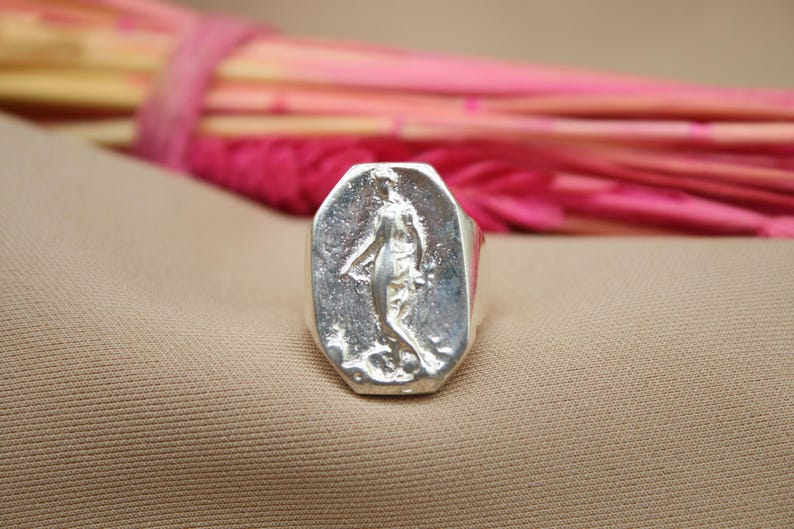 May include: A silver ring with a rectangular face featuring a sculpted figure. The ring is set against a neutral background with a blurred arrangement of pink and yellow stems in the background.