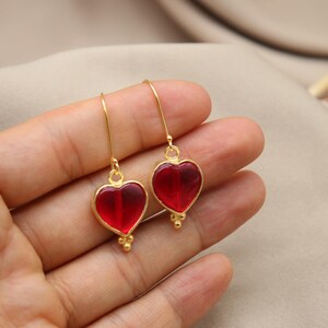 May include: A pair of gold heart-shaped earrings with red glass inserts. The earrings are dangling and have a small gold bead detail at the bottom.