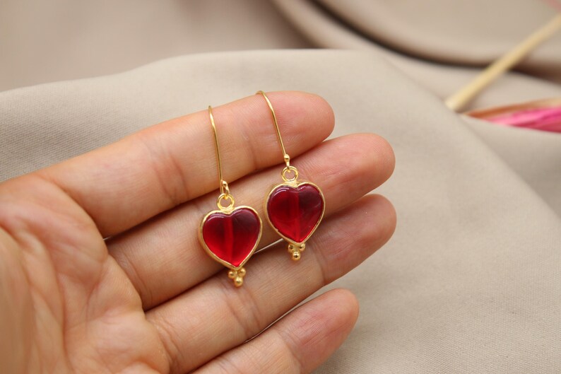 May include: A pair of gold heart-shaped earrings with red glass inserts. The earrings are dangling and have a small gold bead detail at the bottom.