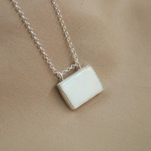 May include: A silver chain necklace featuring a rectangular pendant with a smooth, opaque white inlay. The pendant is securely attached to the delicate silver link chain, which displays a bright, polished metallic finish throughout the piece.