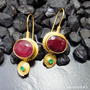 Op de afbeelding: Een paar gouden oorbellen met rode en groene edelstenen. De oorbellen hebben een grote ovale rode edelsteen omgeven door een gouden frame, en een kleinere groene edelsteen gezet in een spiraalvormig gouden frame.