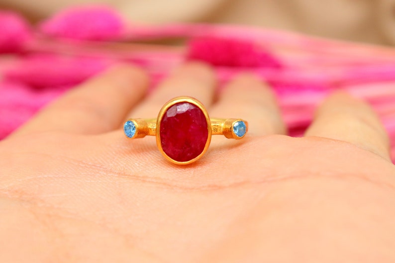 K&ouml;nnte beinhalten: Ein goldener Ring mit einem gro&szlig;en ovalen Rubin-Edelstein und zwei kleineren blauen Edelsteinen auf jeder Seite.