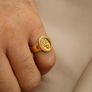 May include: A gold ring with a raised design of a bird on the top. The ring is worn on a person's finger.
