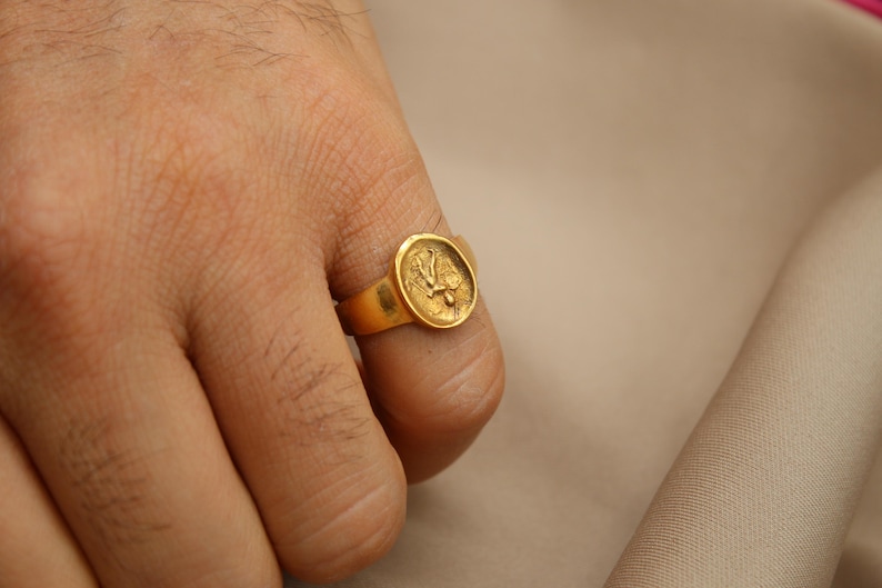 May include: A gold ring with a raised design of a bird on the top. The ring is worn on a person's finger.