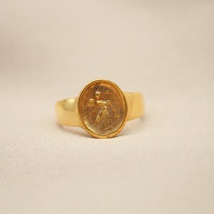 May include: A gold ring with an oval-shaped setting featuring a raised design of a person holding a staff.