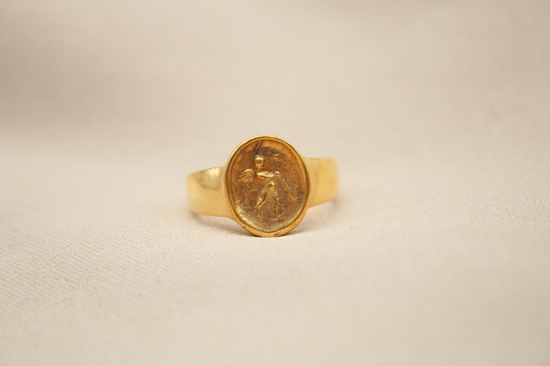 May include: A gold ring with an oval-shaped setting featuring a raised design of a person holding a staff.