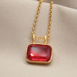 Könnte beinhalten: Eine goldfarbene Halskette mit einem rechteckigen, rubinroten Anhänger. Der Anhänger ist in einen goldenen Rahmen gefasst und hängt an einer zarten Goldkette. Die Halskette wird auf einem neutralfarbenen Stoff präsentiert.