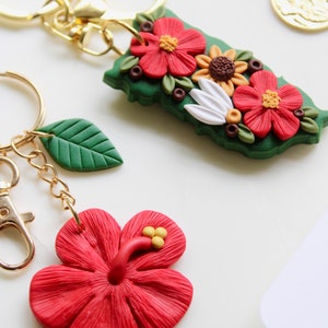 Puede incluir: Dos llaveros con dijes de arcilla polimérica. Un llavero presenta una hoja verde y una flor de hibisco roja. El otro llavero presenta una hoja verde con flores rojas, blancas y amarillas.