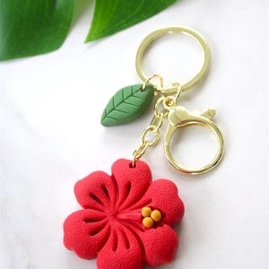 以下が含まれることがあります： 中央が黄色の質感のある赤いハイビスカスの花のチャームと、小さな緑の葉のチャームが付いたゴールドトーンの金属製キーホルダー。花と葉は、短いゴールドの金属チェーンでゴールドの二重リングとロブスタークラスプに取り付けられています。