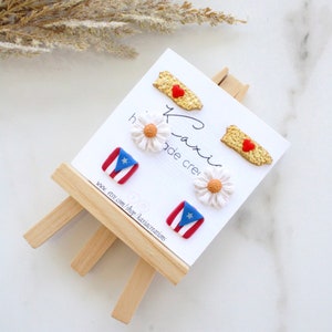 May include: A set of three pairs of earrings featuring a white daisy, a red heart on a gold Puerto Rico outline, and a red, white, and blue Puerto Rican flag. The earrings are displayed on a small wooden easel with the text "Kaxix Creations" and a website address.