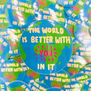 May include: A collection of round, blue and green stickers. Each sticker features a world map design with the text "THE WORLD IS BETTER WITH YOU IN IT" in white and pink. The stickers are scattered across a surface.
