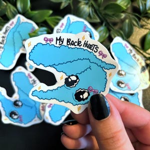 Könnte beinhalten: Blauer Comic-Charakter mit einem flauschigen Körper und großen Augen. Der Charakter trägt eine rosa Schleife und der Text "My Back Hurts" ist darauf geschrieben.