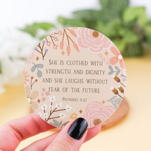 May include: A round sticker with a floral design in shades of pink, white, and green. The sticker features the quote "She is clothed with strength and dignity and she laughs without fear of the future." and the text "Proverbs 31:25".