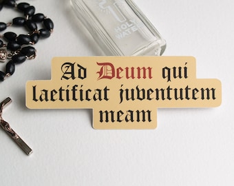 Puede incluir: Una pegatina con la frase latina "Ad Deum qui laetificat juventutem meam" en texto negro sobre fondo beige. El texto está en un estilo de fuente medieval. La pegatina está junto a un rosario negro y una pequeña botella de vidrio etiquetada como "Agua bendita".