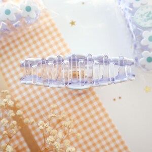 May include: A clear and lavender-colored claw hair clip with a scalloped edge. The clip has a series of vertical bars for gripping hair. The background includes a gingham pattern and small flower-shaped hair accessories.