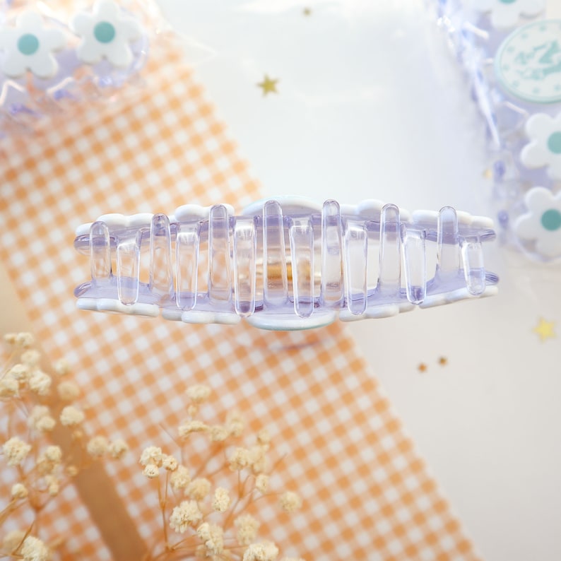 May include: A clear and lavender-colored claw hair clip with a scalloped edge. The clip has a series of vertical bars for gripping hair. The background includes a gingham pattern and small flower-shaped hair accessories.