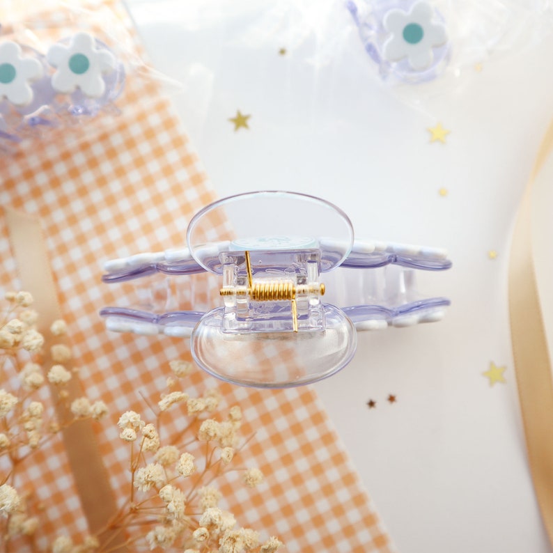 May include: A clear plastic hair claw clip with a gold-colored spring and lavender flower accents. The clip is designed with a scalloped edge and is suitable for various hair styles. Additional flower-shaped hair accessories are visible in the background.
